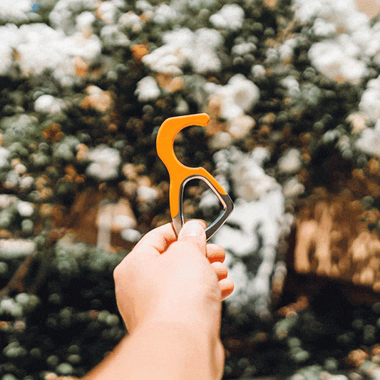 Orange silicone no-touch keychain tool and metal ring, held above bushes